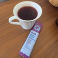 A white mug containing dark red tea sits on a wooden table next to a purple and white tube of Pura Night Time Restore powder containing sour cherry and chamomile