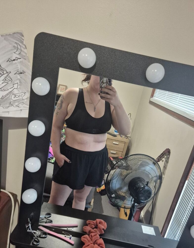 Person taking a mirror selfie in a room with a vanity mirror that has light bulbs around the frame