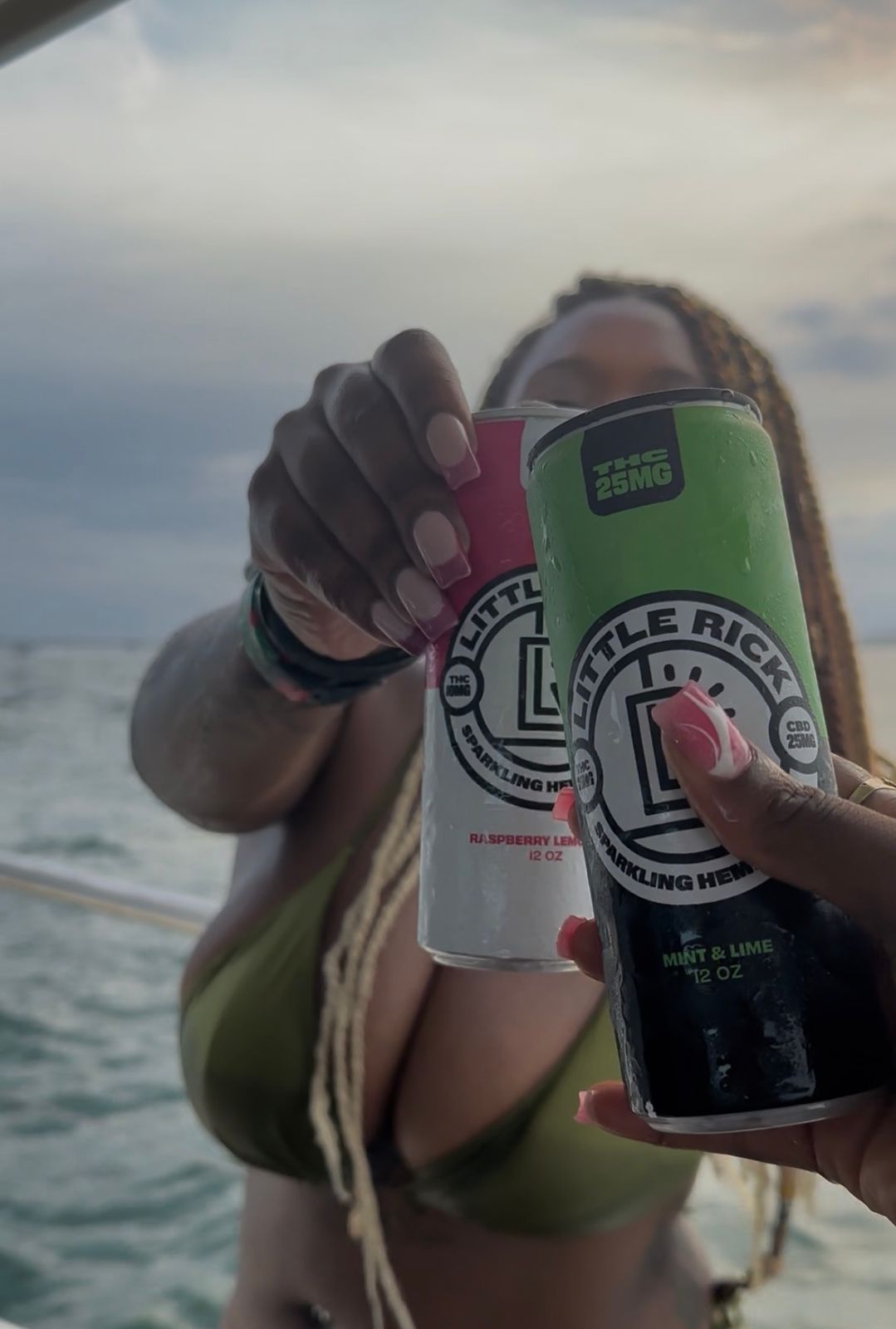 Person holding two cans of Little Rick sparkling hemp beverages in raspberry lemon and mint lime flavors while on a boat with water and sky in the background