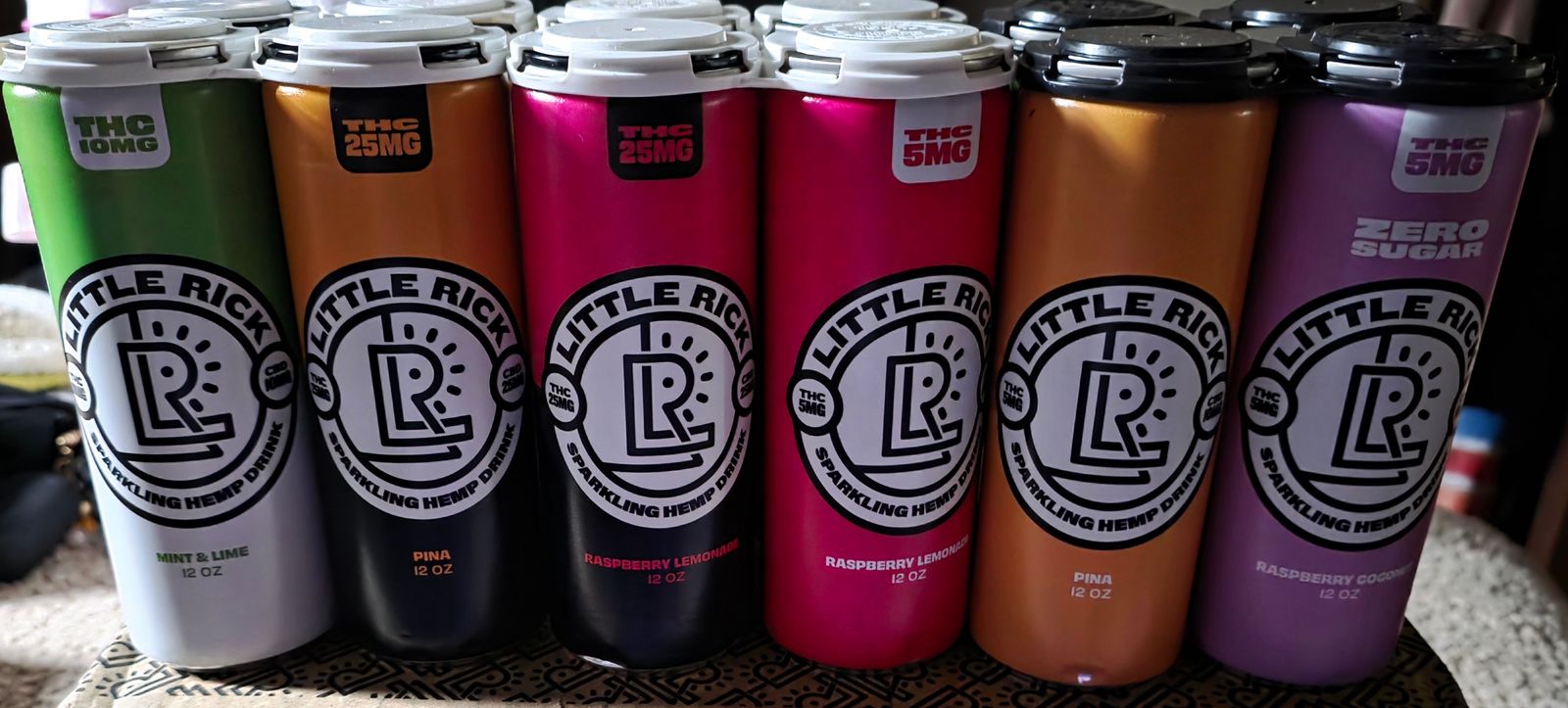 A row of colorful beverage cans with "Little Rick" branding in various flavors including mint lime, pina, and raspberry lemonade displayed on a wire shelf