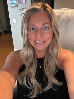 Customer-uploaded image. Click to see more details. The image represents: A woman with long blonde hair smiling at the camera in a home interior setting, wearing a black sleeveless top and a pendant necklace.