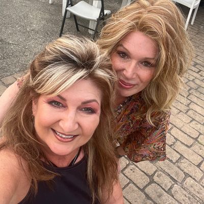 Customer-uploaded image. Click to see more details. The image represents: Two women smiling at the camera outdoors on a paved patio area with white tables and chairs visible in the background.