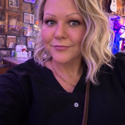 Customer-uploaded image. Click to see more details. The image represents: A woman with shoulder-length blonde wavy hair wearing a black cardigan and gold chain necklace, photographed indoors with purple and warm lighting in the background.