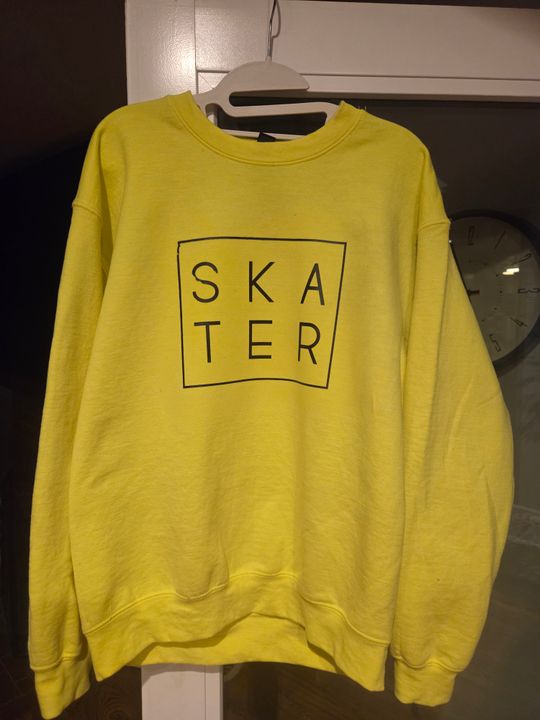 Yellow sweatshirt with SKATER text in a box printed on the front, hanging on a white hanger against a white wall with a clock visible in the background.