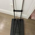 A black folding hand truck or dolly with an extended telescoping handle and flat platform base, positioned on a wooden floor against white cabinet doors