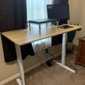 A height-adjustable standing desk with a laptop on a monitor stand, positioned near a window with curtains in a home office setting