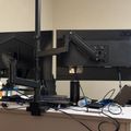 A desk setup with two computer monitors mounted on an adjustable arm, along with various office supplies, cables, and a laptop on a white desk surface against a beige wall.