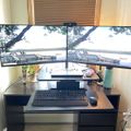A home office setup with dual monitors displaying a lakeside scene, positioned on a dark wooden desk with keyboard and mouse, near a window with wooden blinds
