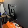 A 3D printer extruder head mounted on a black bracket against a gray wall, with an orange build platform visible below and a hand reaching toward the device.