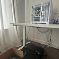 A white gaming PC with transparent side panel sits on a white desk next to a window with white curtains, with various cables and a cardboard box visible below the desk.