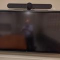 Wall-mounted TV with video conferencing camera attached on top, displaying a blurred screen reflection
