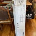 A long white product box for a wall mount or mounting system is leaning against a wooden dining table in a home setting, with an Adidas bag visible on a chair nearby.