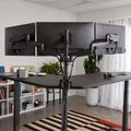 Modern office workspace with three black monitors mounted on adjustable arms above a dark desk, featuring white bookshelves, plants, and a striped area rug