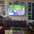 Living room with large wall-mounted TV showing animated content, wooden entertainment center below, coffee table with glass top, and various framed pictures on walls