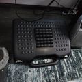 Black ergonomic foot rest with massage bumps and adjustable height positioned under a desk on a patterned carpet