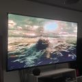 Large TV screen displaying a video game scene with a boat on water, mounted on a white entertainment stand in a dark room