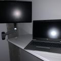 A laptop computer sits on a white desk next to a wall-mounted monitor on an adjustable arm in a modern office setup