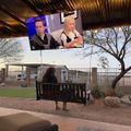 Outdoor patio area with covered seating, landscaping, and a large TV screen displaying two people in what appears to be a news or talk show program
