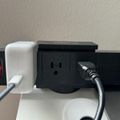 A black power strip with multiple outlets and USB ports, with various cables and chargers plugged in, sitting on a white surface