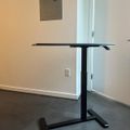 A height-adjustable standing desk with black metal frame and dark desktop surface positioned in a room with white walls and gray flooring