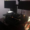 A dual monitor computer workstation setup on a dark desk with an adjustable standing desk converter, located in a room with purple walls