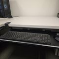 A computer desk setup with a keyboard and mouse on a pull-out tray, with a computer tower and other equipment visible on the white desk surface above
