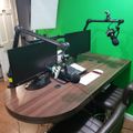 A home studio setup with a curved wooden desk, two black leather chairs, computer monitors, microphone arms, and a bright green wall background used for video recording or streaming.