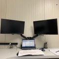 A white corner desk workspace with a laptop computer, dual monitor arm setup with two black monitors, papers, and office supplies against white paneled walls