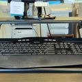 A workspace setup showing a Logitech keyboard in the foreground with a Samsung monitor elevated on a stand in the background, along with various office supplies and sticky notes on a wooden desk surface.