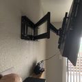 A black TV wall mount bracket extended from a white wall in a corner, with a television attached and various electronic devices visible on a shelf below