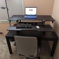 A home office setup with a black standing desk converter, laptop, keyboard, mouse, desk lamp, and white office chair positioned near a window with blinds