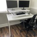 A clean white desk setup with dual monitors, an adjustable standing desk converter, keyboard, mouse, and black ergonomic office chair on hardwood flooring in a corner office space