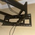 A black adjustable TV wall mount bracket installed on a beige ceiling, with extending arms and cable management visible
