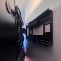 Two black TV wall mounts installed on opposite walls in a narrow hallway, with blue and red LED light strips running along the gap between them, illuminating the white textured ceiling above.