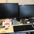 A dual monitor computer setup on a desk with keyboard, mouse, and various office supplies scattered around