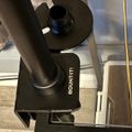 Black telescope mount with threaded connector and adjustment knob, labeled "MOUNT", attached to a desk or table edge with wooden furniture visible in the background.