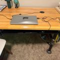 A wooden desk with adjustable black legs holding a closed laptop computer with various cables connected to it