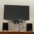 Wall-mounted flat screen TV above a wooden cabinet with speakers and electronic devices on a glass shelf