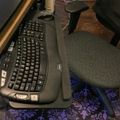 A black ergonomic keyboard sits on a desk next to a computer monitor, with an office chair and decorative purple patterned rug visible in the workspace