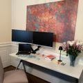 A modern home office setup with dual monitors, laptop, white desk, beige chair, and decorative flowers, featuring a colorful abstract artwork on the wall