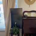 A cozy room corner with beige curtains, a dark wooden dresser with mirror, a straw hat hanging on the wall, a potted plant in a gold planter, and a black TV screen near a window with blinds.
