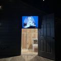 Dark gallery space with wooden platform leading to illuminated doorway and blue video screen displaying a figure