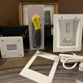 Various electronic device mounting plates and wall outlets displayed on a wooden surface, including white rectangular wall plates with openings and a packaged tool with yellow handle