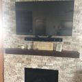 A flat-screen TV mounted above a stone fireplace with a dark wooden mantel shelf containing decorative items