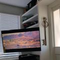 A home office or living space with a large monitor displaying a desert landscape scene, white built-in shelving, window blinds, and a decorative wall hook near a white door.