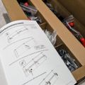 An open cardboard box containing packaged items with red caps or components, alongside installation instruction sheets with technical diagrams showing assembly steps.