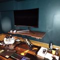 A home office workspace with a curved monitor on an adjustable wooden desk, gaming keyboard, VR headset, and various office supplies against teal painted walls