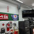 Interior of an automotive garage or workshop with white walls, fluorescent lighting, a mounted TV screen, various automotive equipment and tools, safety equipment including a fire extinguisher, and automotive brand signage on the walls.