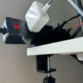 A power strip with multiple plugs and cables attached, mounted under a white desk or shelf with a black clamp mechanism