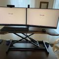 Dual monitor computer workstation setup on an adjustable standing desk with keyboard, mouse, and webcam in a home office environment